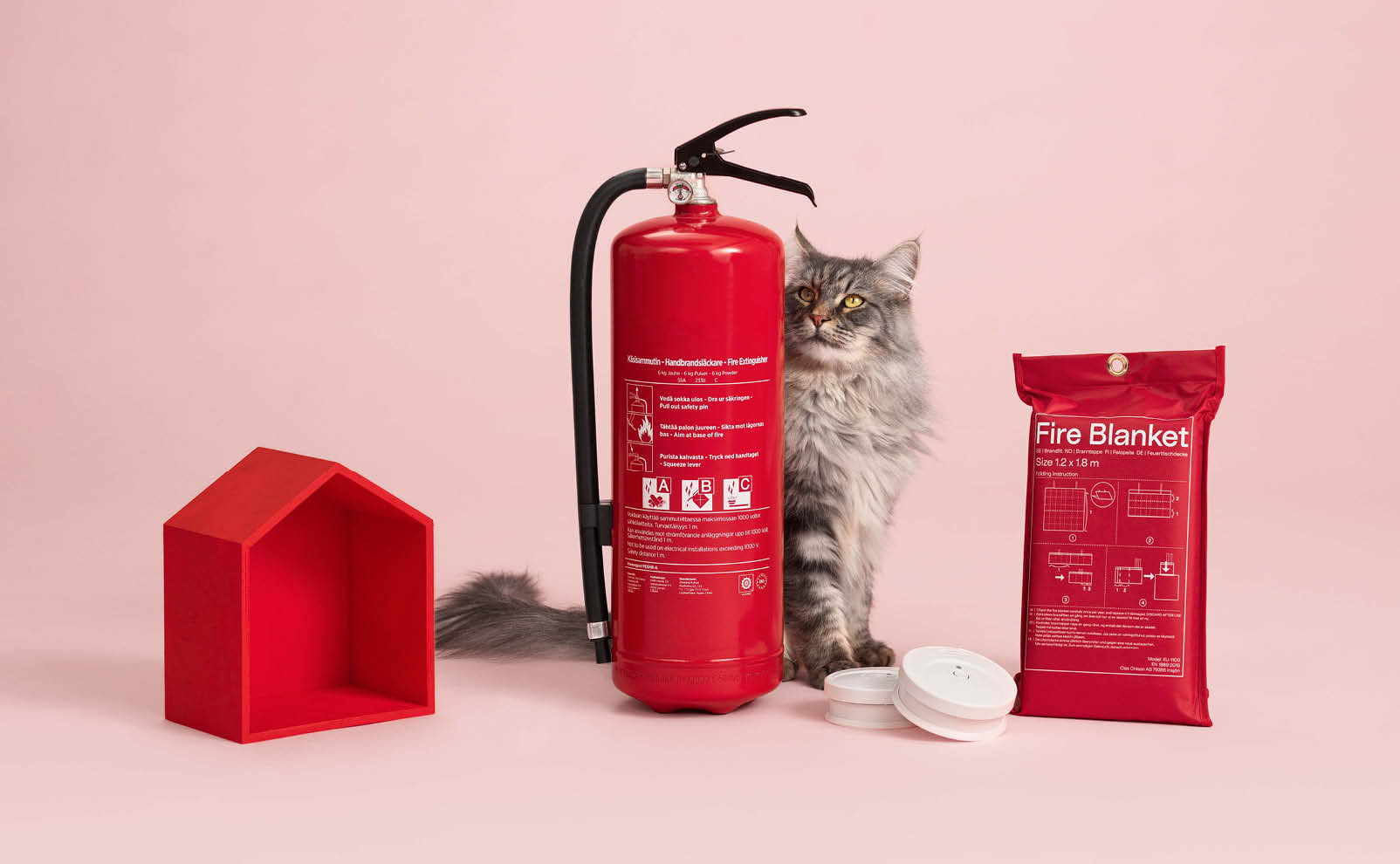Katt sitter bakom en brandsläckare och brandvarnare. Har en brandfilt och ett hus bredvid sig. 